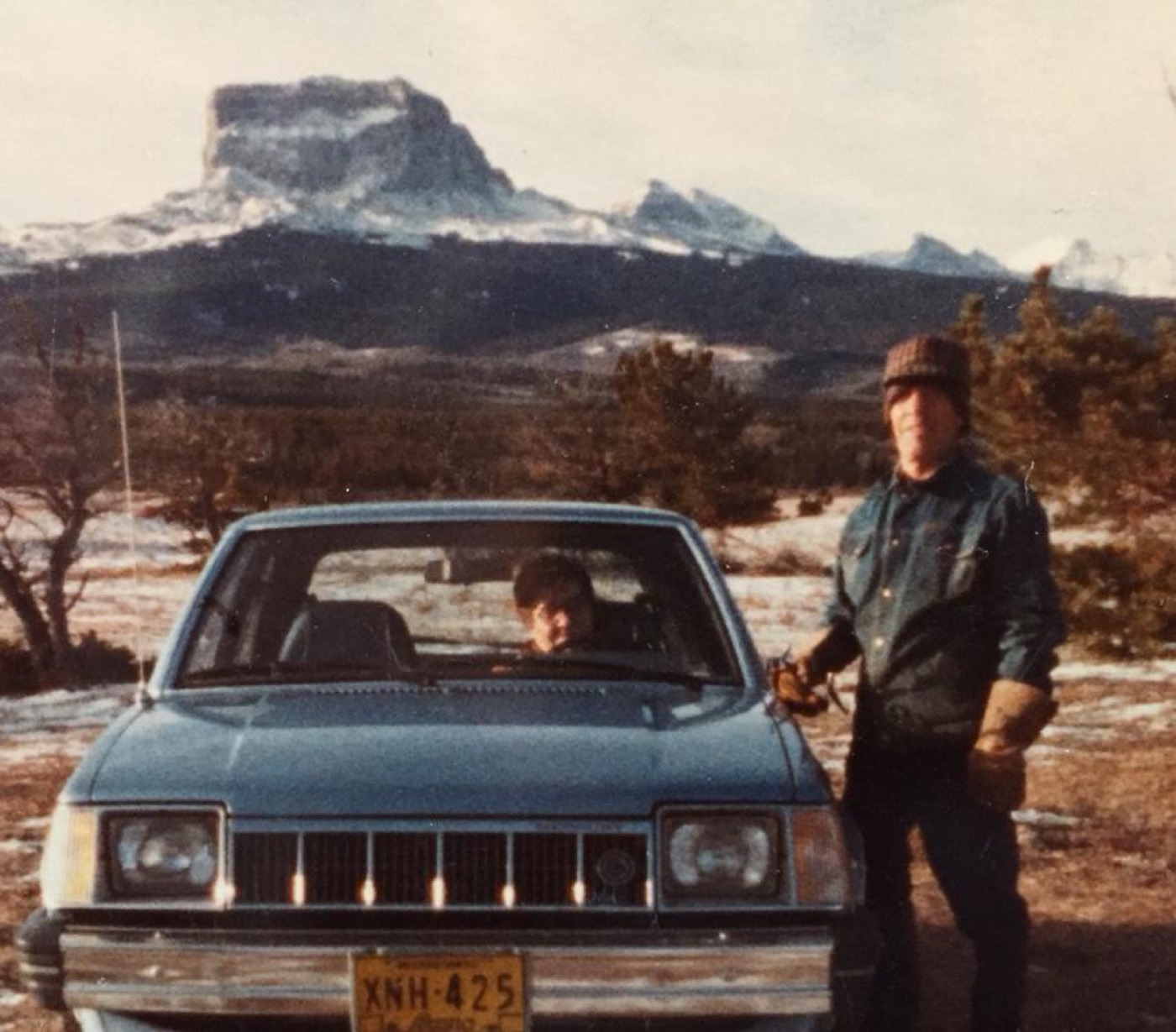 A faded film photograph of a man and a boy beside a blue car, with Chief Mountain behind them
